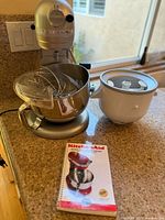 Front view of mixer assembled with bowl, wire whisk, pouring shield, instruction manual and ice cream attachment placed beside it