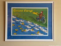 Full front view of framed artwork showing Curious George on bicycle riding alongside river filled with paper boats and rubber ducks