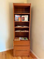 Front view of tall teak bookshelf with three open shelves and two lower drawers on hardwood floor against wall.