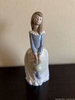 Front angle view of the figurine depicting a seated girl holding a hat in front of her.