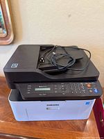 Samsung Xpress M2070FW printer scanner copier with power cord on top, shown from front angle.