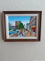 Front view of framed painting showing Paris city scene with Notre Dame cathedral, river, and people walking on cobblestone sidewalk.