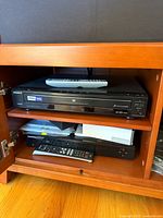 Two black DVD players stacked inside a brown wooden TV cabinet shelf. The top device is a Sony DVD player with an HDMI badge and a remote on top. The bottom device is a Samsung DVD recorder with a remote resting in front of it.