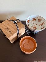 Photo showing the porcelain bowl on its wooden stand, small brown ceramic bowl, and wooden box with ribbon on a wood surface