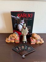 Full view of Asian art set: two wooden hand-painted reclining figure bookends, a standing figurine, a decorative folding fan, and the book 'Kabuki'.
