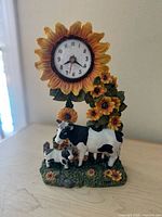 Front view of vintage cow and sunflower clock showing the clock face surrounded by sunflower petals and cow figurines standing on grass with sunflower accents.