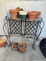 Vintage black wrought iron garden table with lattice top and curved legs, holding eight assorted plant pots of terracotta, ceramic, and plastic materials, some with plants.