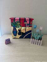 Front view of small hand painted wooden tabletop cabinet, purple patterned cube, and wooden flower decoration.