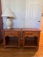 Pair of vintage Ethan Allen knotty pine wooden nightstands shown side by side against a white wall with a white door in the background. A lamp with a white base and beige shade is placed on the left nightstand (lamp not included in sale).