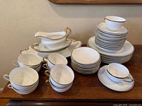 Photo showing multiple white ceramic cups, stacks of saucers and plates, and serving pieces with gold trim accents.