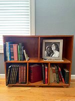 Front view of vintage wooden bookcase against grey wall with window blinds, showing four compartments containing books and framed photograph.