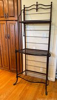 Full view of the vintage wrought iron bakers rack showing its height, three shelves with a wooden top shelf, and decorative finials.