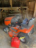 Full view of orange Ariens riding mower with battery on seat and fuel can in foreground
