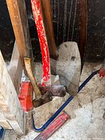 Mixed hand tools leaning in corner showing axe, hammer, spade, red scoop and blue crowbar