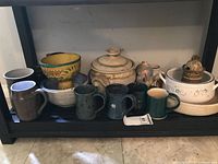 Shelf showing Pampered Chef base, lidded casserole, decorative bowl, small jug and five mugs