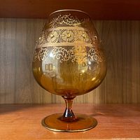 Front view of amber hand blown glass snifter showing gold floral and flourish decoration on bowl and rim with round stem and base