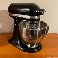 Front-left view of black mixer with stainless steel bowl and wire whip attached