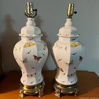 Two white ceramic urn shape table lamps with butterfly patterns, placed side by side showing shiny gold base on left lamp and antique gold base on right lamp. Both have brass bulb sockets, no shades. Shown from front view.