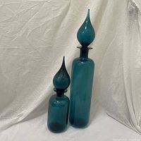 Pair of tall and short celadon blue glass genie bottles against white fabric background.