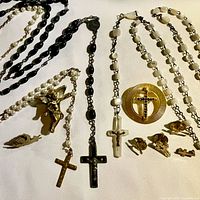 Photo showing three rosaries with varied bead materials and crosses, plus two angel pins and several cross pins on white background.