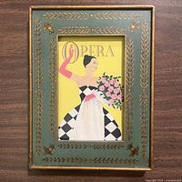 Front full view of framed vintage fashion poster titled 'L’Opera' with woman in checkered dress and pink gloves holding flowers, inside Florentine frame with green and gold leafy design.