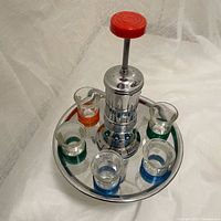 Top-down image of the chrome tray with glass bottle inside and six shot glasses arranged around a chrome pump with red button top.