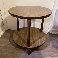 Front angle view of two-tier round side table with walnut veneer tops and chocolate turned legs on dark floor against white wall.