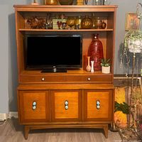 Front view of MCM storage cabinet showing open shelf with decorative items, TV space, two drawers below, and three-door cabinet with turquoise knobs.