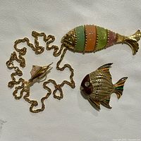 Photo showing the full nautical jewelry set including the reticulated fish necklace with multi-colored carved glass segments, the gold tone fish brooch with enamel details and rhinestone eye, and the gold tone marlin pin.