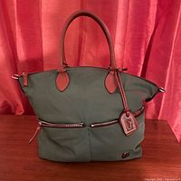 Front view of the green nylon tote bag with brown leather handles and two exterior zippered pockets, displayed on wooden table against red curtain background.