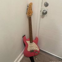 Full view of pink electric guitar leaning against corner showing body, neck, and strap.
