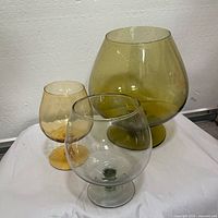 Photo showing three oversized hand blown glass snifters in amber, smoke, and greenish shades on a white cloth background.