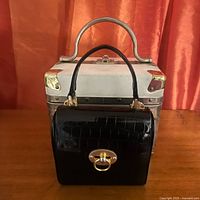 Shows front view of the two purses stacked, with black embossed mini box purse in front of larger white hard case purse with silver corners and handle