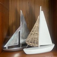 Front and side view showing two sailboat figurines, one hammered aluminum and one painted wood with wicker sail, displayed on a wooden shelf.