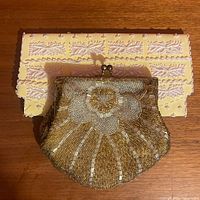 Gold beaded purse with floral pattern in gold and white seed and barrel beads, scalloped bottom, gold-tone frame and chain, placed on yellow purse