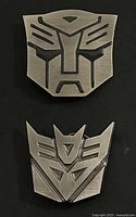 Two metal belt buckles, one with the Autobot logo and the other with the Decepticon logo, both in brushed silver finish with black recessed details.