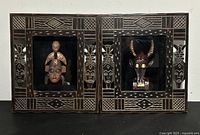 Full view of both framed African tribal mask shadow boxes side by side with carved wooden borders and black fabric background.