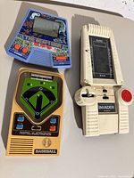 Photo of three handheld electronic games: Casio The Beam War (blue triangular), Mattel Baseball (beige with green baseball diamond), Gakken Invader (white with black screen and red button) showing overall condition and design.
