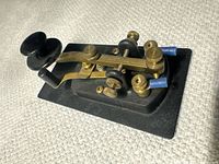 Side and angled view of the Morse code telegraph key showing brass lever, black plastic knob, and base with steel and brass components.