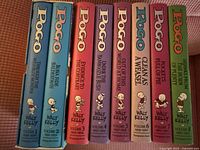 Photo of the 8 Pogo comic volumes showing their spines with volume titles, author Walt Kelly, and vibrant colored covers in good condition.