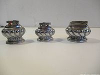 Three chrome-tone metal table lighters lined up showing size variation and design