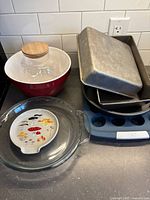 Photo showing red ceramic bowl with clear glass canister that has a wooden lid, mushroom ceramic spoon rest, clear Anchor Hocking pie plate, and multiple aluminum bake pans including Betty Crocker and Bake-Rite tins plus a blue silicone mini muffin tray.