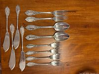 All 12 flatware pieces arranged on table: 4 spreaders at left, 4 dessert forks at bottom right, 2 dinner forks at top right, 2 serving spoons in middle