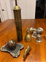Photo showing the tall brass container, decorative brass bell, four small silver-colored bells, round perforated brass container with ornate base, and brass crocodile figurine arranged on a wooden surface