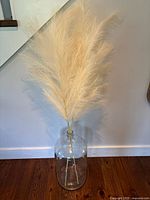 Full view of large clear glass carboy with tall faux pampas grass stems arranged inside, displayed on wood floor against wall.