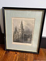 Framed etching print showing the full image of the cathedral from a front-angle view, placed on wooden floor against a white wall.