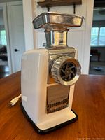 Side view of the Grind-O-Matic electric grinder showing white body, chrome feed chute and grinding plate, black rubber base.