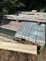 Photo showing stacked corrugated and flat metal sheeting with some wood planks on top outdoors.