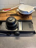 Photo of Cuisinart mandolin slicer on counter showing adjustable knob and black safety holder, wooden cutting board, mixing bowls and utensils in background.