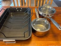 Photo showing both stainless steel pots with glass lids alongside the black roasting pan with removable rack on a wooden table.
